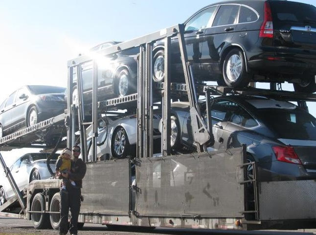A driver holding his baby son in front of a car carrier loaded with vehicles.